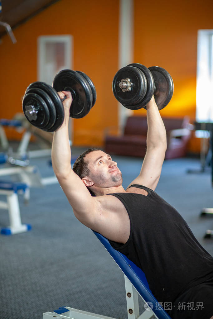 在体育俱乐部举重的强壮男子在健身房用哑铃练习肌肉帅气的健身教练在