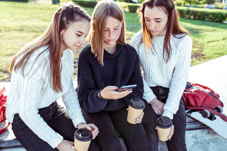 三名1214岁的女学生，夏天在城里，手里拿着智能手机，喝茶喝茶，在网上看照片。周末休息。休闲服，保暖毛衣。休学大学。