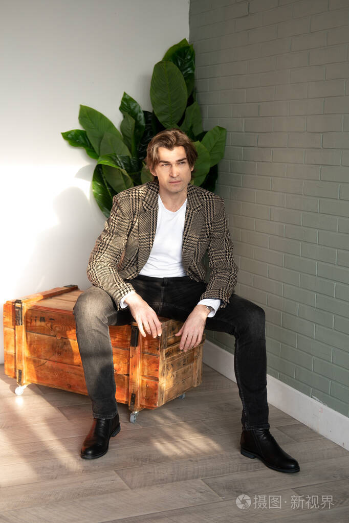 高大英俊的男人坐在木箱上旁边是绿色的植物和灰色的墙壁