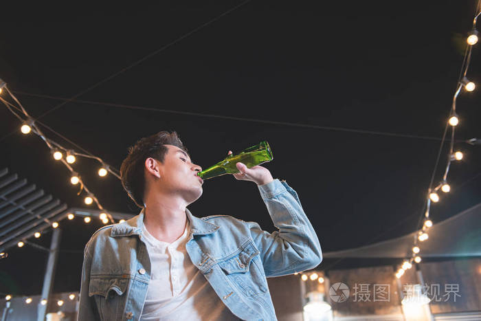 帅气的时髦男人坐在餐厅夜总会派对屋顶层的吧台前,手拿啤酒瓶喝酒的