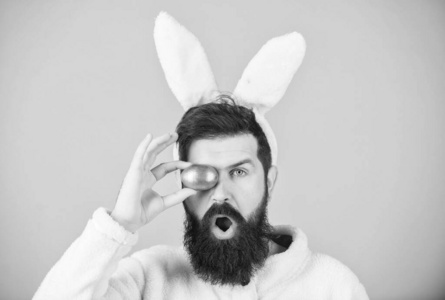 长着胡子和胡子的滑稽兔子拿着粉红色的鸡蛋。胡子男人戴着愚蠢的兔子耳朵。复活节象征概念。我的宝贝。时髦可爱的兔子长耳朵蓝色背景。复