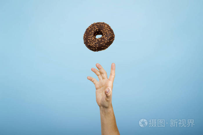 用手抓一个涂有巧克力糖霜的甜甜圈蓝色硬纸板背景烘焙概念手工制作平