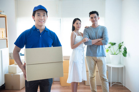 微笑着的年轻亚洲送货员穿着蓝色制服，拿着两个名片盒包裹看着相机，快乐的亚洲年轻夫妇的寄件人站在背景中。