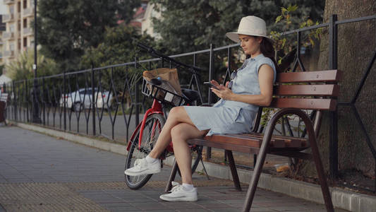 一个戴着帽子和衣服的女人坐在一辆红色自行车旁边的长凳上，用她的电话。