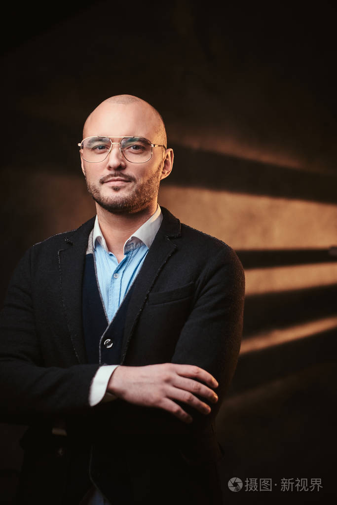 一个英俊的光头商人穿着黑色夹克蓝色衬衫戴着眼镜在黑暗的工作室里摆