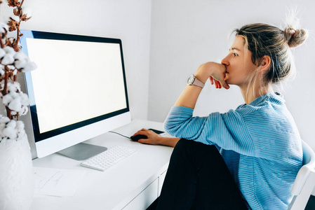 自由职业女商人在办公室使用台式电脑的侧视图。年轻的金发女郎坐在家里，在电脑前工作，屏幕上显示着你的短信或内容。