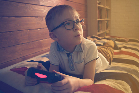 戴眼镜的白人男孩躺在他房间的床上，手里拿着一根操纵杆。他在控制台上玩电子游戏