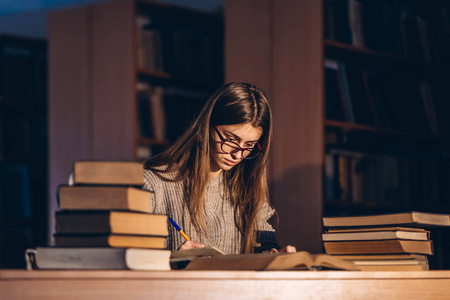 戴眼镜的年轻学生准备考试。晚上的女孩拿着一堆书坐在图书馆的桌子旁