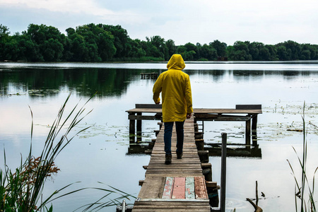 穿着黄色防水雨衣的时髦年轻人站在湖边或河边的木码头上照片