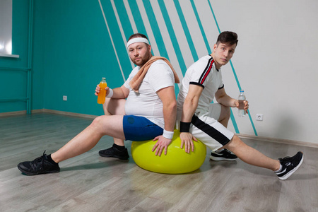 胖子和他的私人教练坐在健身球上，拿着几瓶水在健身班上辛苦训练。那个超重的男人非常疲倦