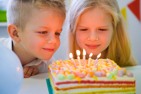 两个金发碧眼的白人男孩和女孩看着生日彩虹蛋糕，点燃蜡烛，并在生日聚会上许愿。彩色背景