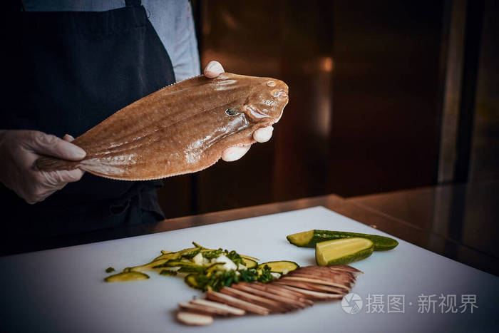 厨师站在一个黑暗的餐厅厨房里旁边放着蔬菜手里拿着一条大鱼围着围裙
