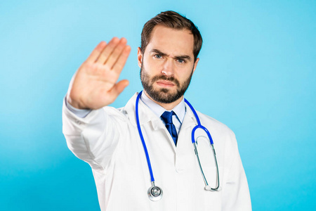 身着专业医疗白色外套的严肃医生的肖像，通过停止手势显示拒绝手势。医生被隔离在蓝色背景下。