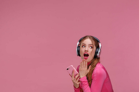 奇怪的红头发女孩拿着手机等待朋友的留言和祝贺。戴着耳机的微笑的年轻女子拿着粉红色背景的智能手机。复制空间