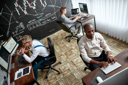 三名交易员在办公室工作时,坐在电脑显示器前的桌子旁的高角度视图.