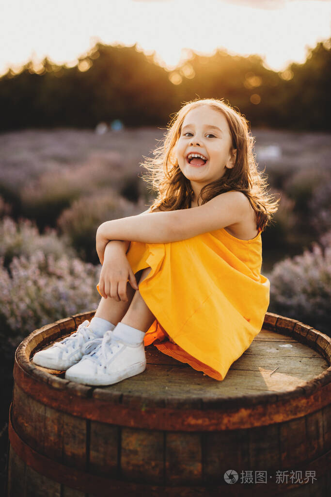 穿着黄色连衣裙的白人小女孩坐在薰衣草地上的桶上开心地笑着