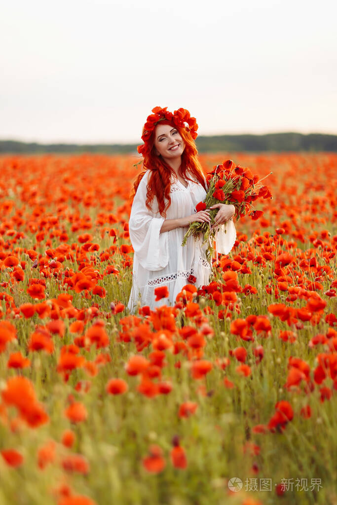 美丽的红发微笑的女人在绿色的田野上戴着花环开着一束罂粟花