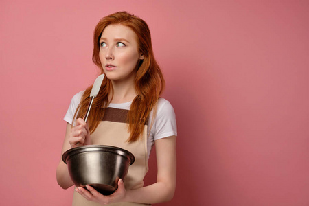 一个穿着围裙和T恤的红发女孩站在粉红色的背景上，手里拿着碗和搅拌器，看起来很害怕