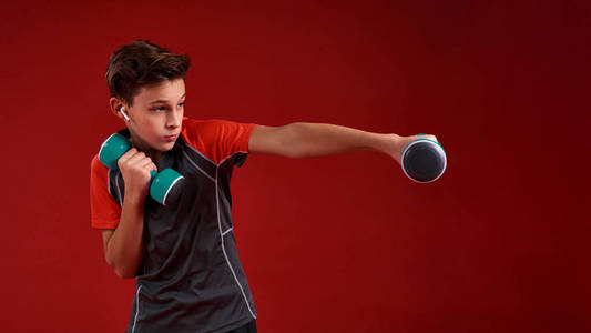 力量。一个十几岁的男孩从事体育运动，一边用哑铃一边看一边。隔离在红色背景上。训练健身积极的生活方式理念。水平射击