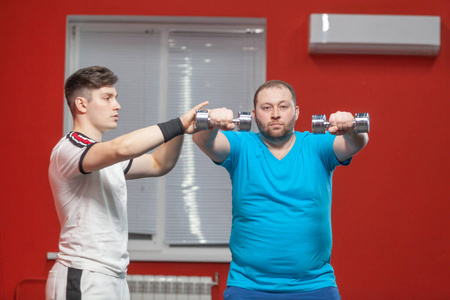 私人健身教练向一个胖子展示如何在健身房用哑铃进行锻炼。超重