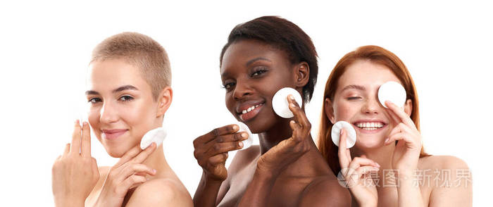 所有人都需要护肤品。三位积极多元文化的年轻女性在白色背景下站在工作室里，用棉垫卸妆，微笑着