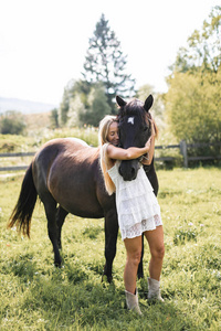 美丽的年轻金发女子，穿着白色波西米亚裙，在日落时亲吻和拥抱她的马，夏日野外场景