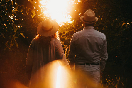 一对戴着漂亮帽子的中年白人夫妇在一个满是树木的花园里散步，一边看着夕阳