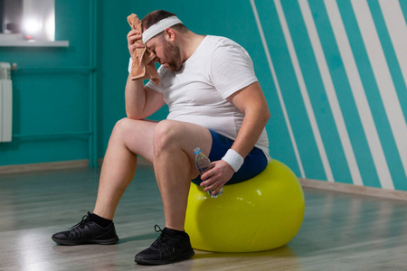 胖子用毛巾擦额头上的汗在集体健身课上,超重男子躺在健身球上.