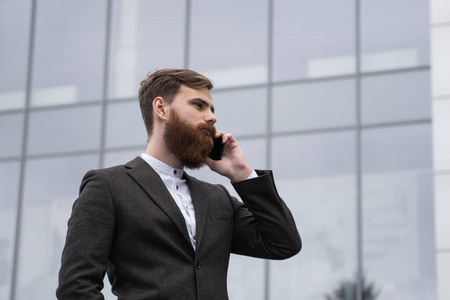 在金融区打电话的商人。一位年轻的城市职业男子在办公楼上空使用智能手机的肖像。在户外用智能手机说话的商人。技术。