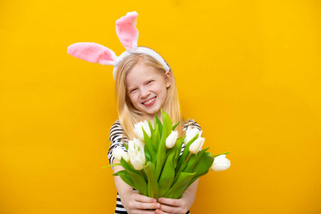 黄色摄影棚背景上，头戴兔子耳朵微笑的女孩。快乐的疯狂的快乐的孩子和郁金香花束。复活节和春天