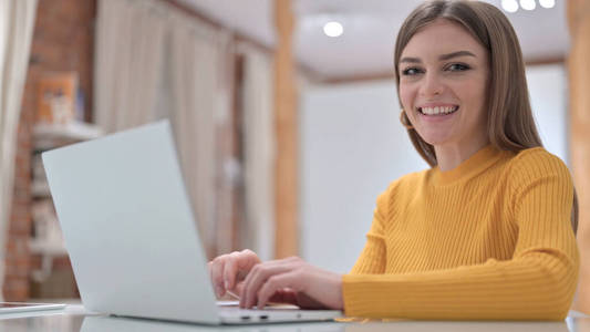 有创意的年轻女士在笔记本电脑前工作，对着镜头微笑