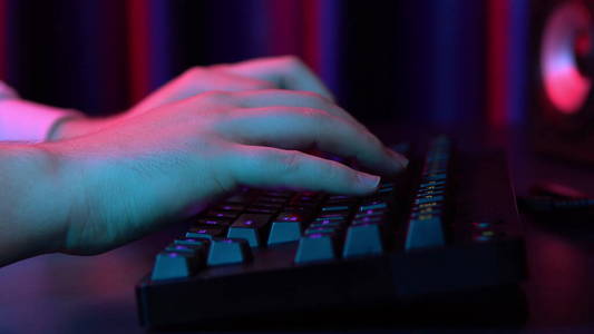 一个年轻人正在电脑键盘上打字。手放紧。蓝色和红色的光照在手上。