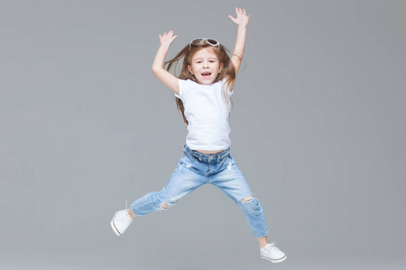 裤白色t恤和太阳镜的学龄前儿童在灰色背景下孤立地举起双手跳跃照片