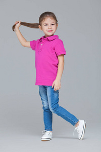 一个穿着休闲服的活泼有趣的小女孩的全幅肖像牛仔裤和粉色t恤，一只手拿着马尾辫，在灰色的工作室背景下