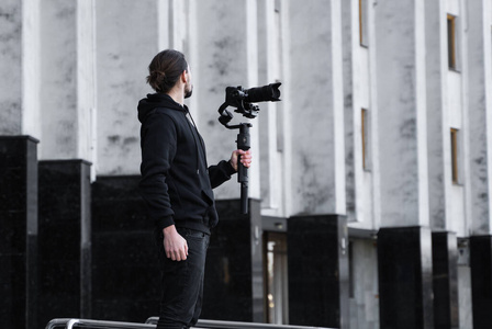 年轻的专业摄像师在3轴万向节稳定器上手持专业摄像机。专业设备有助于制作高质量的视频而不震动。摄影师穿着黑色连帽衫拍摄视频。