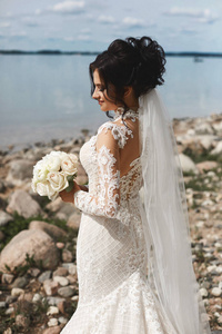 戴着白色蕾丝礼服的婚礼发型的年轻新娘站在湖边。春天，一个穿着婚纱，手持一束鲜花的年轻女子在湖边摆姿势。婚礼时尚