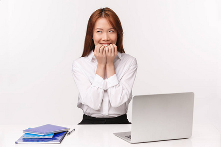 职业工作和女企业家概念。照片中狡猾而体贴，梦幻般傻傻的亚洲女孩坐在办公桌旁，拿着文件和笔记本电脑，心事重重，咯咯笑着