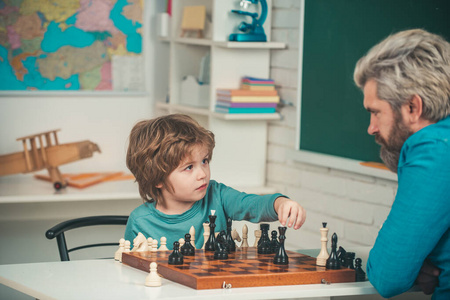 专注的小男孩父子俩坐在桌边下棋。父亲在家教学象棋的男孩。儿童早期发育。