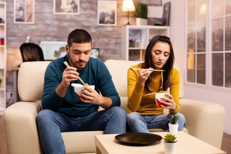 在现代舒适的客厅里，夫妻俩一边看电视一边吃外卖面条