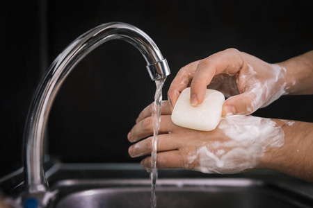 一个年轻人在水龙头下用肥皂洗手，在黑色背景的金属水槽上方。病毒预防。卫生程序。洗手。