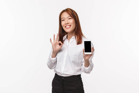 这很好。满意漂亮的亚洲女性展示手机显示和做出好的手势，评价优秀的应用程序，为拍摄酷照片感到自豪，站在白色背景下