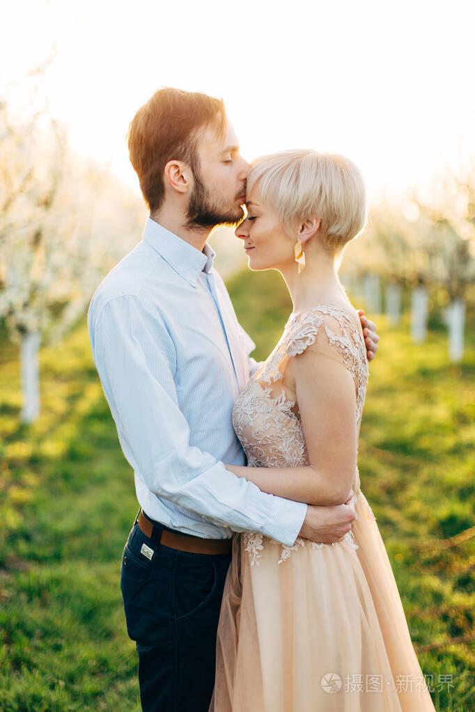 快乐的年轻男子新郎站在鲜花盛开的花园里拥抱他的女新娘亲吻她的额头