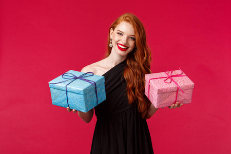 庆祝节日和女性观念。画像中优雅华丽的年轻红发女子身着黑色派对礼服，手持蓝色和粉色两个盒子里的礼物，微笑的相机欢快，红色背景