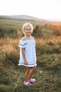一个金发的白人小女孩穿着浅蓝色的裙子，在一片盛开野花和野草的夏日田野里，微笑着看着镜头