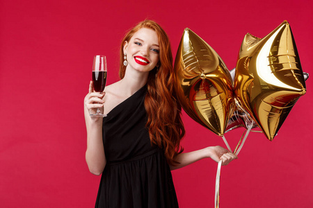 美丽的年轻优雅女人，用红唇膏，姜发和黑色礼服，捧着气球和香槟，为生日快乐的朋友祝酒。