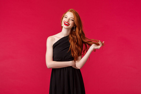 优雅时尚女性理念。自信性感时尚的年轻红头发女人红唇膏，晚妆，穿着优雅的黑色礼服，在正式场合大笑，红色背景