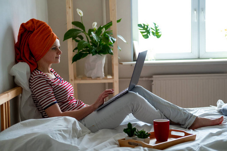 头上戴着橙色毛巾的女人在床上远程办公。使用计算机。远程教育在线教育和工作。