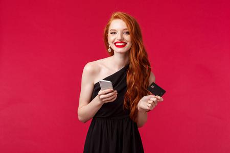 迷人的年轻红发女孩的肖像，穿着黑色连衣裙，长着姜黄色的头发，手持信用卡和智能手机，在线购物，为派对订购新衣服，红色背景