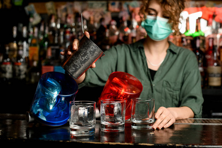 戴着医用面罩的调酒师女孩开始把饮料从钢杯里倒进加冰的玻璃杯里。