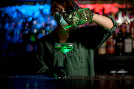 年轻的调酒师女孩戴着医用面罩和黑手套，准确地将绿色饮料从玻璃杯中倒入酒杯。